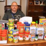 Fulton Rotary President Charlie Anderson sits behind his desk covered with some of the 100 pounds of food donated by the club to the SERVE food pantry. The club also collected $2,170.
